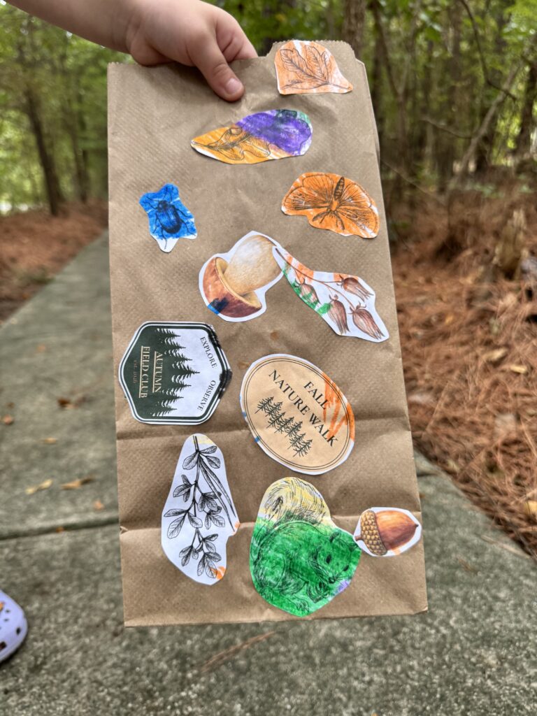 A child holding a finished brown paper nature treasure bag decorated with colorful leaves, a squirrel, botanical cutouts, and autumn field club labels.